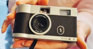 A person holds a vintage-style YesiStar S1 camera with a 31mm f/11 lens. The camera has a silver and black body with a textured grip, and a flash symbol on the front. The camera is centered in the image with the person’s fingers visible at the edges.