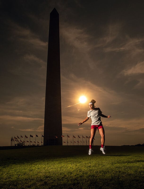 A person wearing a white shirt and red shorts is jumping in the air with arms spread wide in front of the Washington Monument at sunset. The sky is slightly cloudy, and a burst of sunlight illuminates the scene from behind the monument.