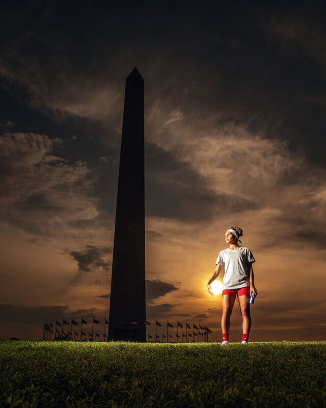 A person dressed in a white t-shirt and red shorts stands on grass with the Washington Monument in the background. The sky is filled with dramatic clouds illuminated by the setting sun, casting a warm glow over the scene. The person holds a drink bottle.