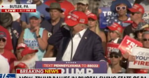 A crowd gathers around a podium with "Trump" signage at a rally in Butler, PA. Many attendees wear red caps and hold signs. A man at the podium, wearing a red cap, speaks into a microphone. News ticker below reads, "BREAKING NEWS NOW: TRUMP RALLIES IN PIVOTAL SWING STATE OF PA."  .