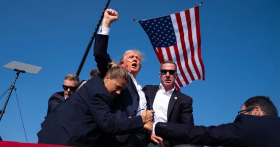 A man in a suit with his fist raised stands among security personnel with earpieces and sunglasses. An American flag waves in the background against a clear blue sky. The scene appears to be outdoors.