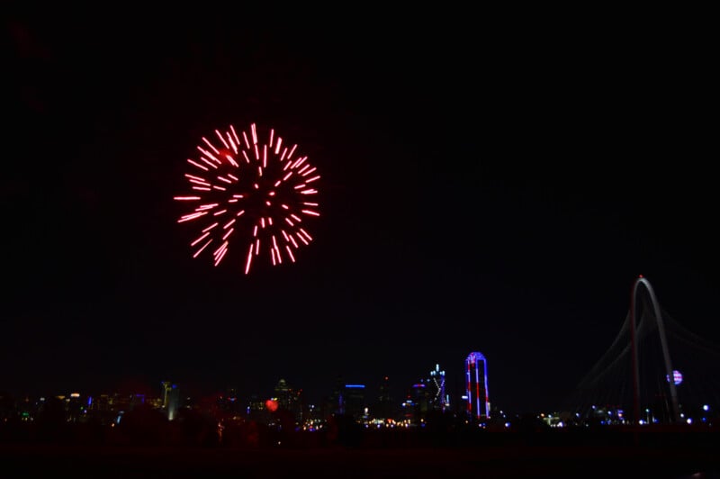 A night show with red fireworks exploding in the sky.  Below, a beautiful view of the city can be seen, including a unique bridge with a long skeleton.  The cityscape has a variety of bright and colorful buildings.