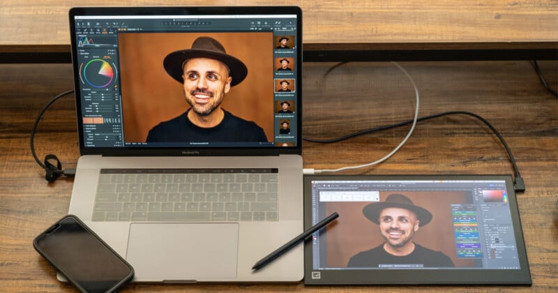 A laptop displaying a photo editing software with an image of a smiling man wearing a brown hat. A tablet connected to the laptop also shows the same image, with a stylus placed on it. A smartphone lies beside the laptop on a wooden table.