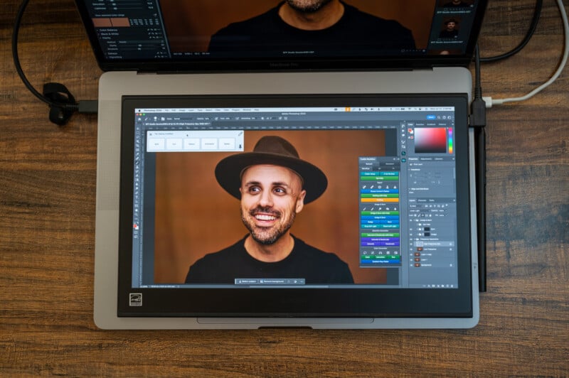 A laptop displaying a photo-editing software with an image of a smiling man wearing a hat. The laptop is on a wooden surface, and another screen is partially visible above it, showing the same image of the man. Various editing tools and panels are open on the screen.