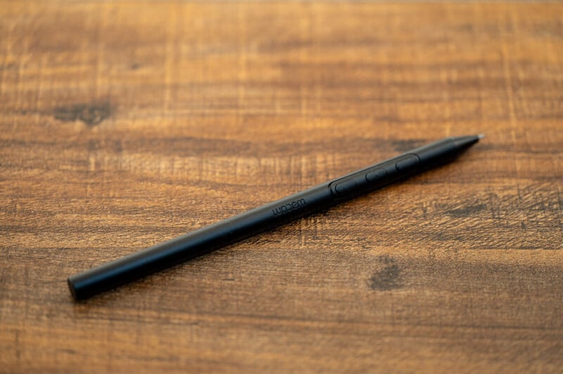 A black stylus pen rests on a wooden surface, showing its side with buttons and the inscription "Wacom" visible. The surface has a warm, brown tone with noticeable grain patterns. The pen is sleek and modern in design.