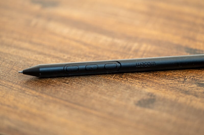 Close-up of a black Wacom stylus pen resting on a wooden surface. The pen is angled diagonally, and three buttons on the side of the stylus are visible. The surface has a warm tone with visible wood grain texture.