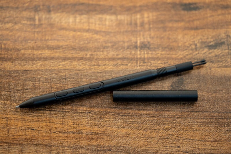 Image of a black Wacom stylus pen on a wooden surface with a cap removed and placed next to it. The pen has two buttons near the tip and features fine detailing with visible branding on the body.