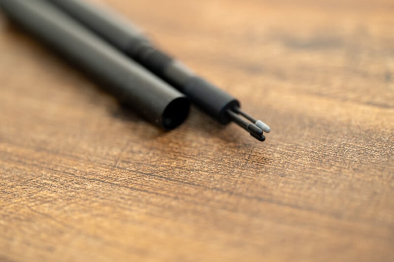 Close-up image of a mechanical pencil with its cap removed, showing two pieces of lead sticking out from the tip. The pencil is laying on a textured wooden surface, with the focus on the lead and the pencil's tip, leaving the background slightly blurred.