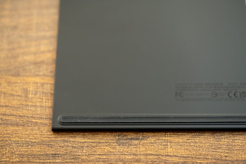 Close-up view of the corner of a sleek, black electronic device, possibly a tablet or laptop, resting on a wooden surface. The corner shows a small engraved text including certification logos such as FCC, CE, and UKCA.