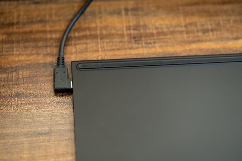 Close-up image of a corner of a black electronic device plugged in with a power cord on a wooden surface. The plugged-in corner is likely the device's charging port. The background shows the texture of the wooden surface.