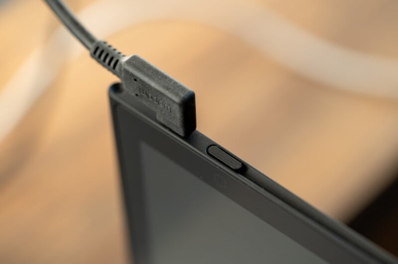 Close-up of a heated display cable plugged into a monitor. The background appears to be a wooden surface, slightly out of focus. The setup suggests a connection between the display and a power source or another device.