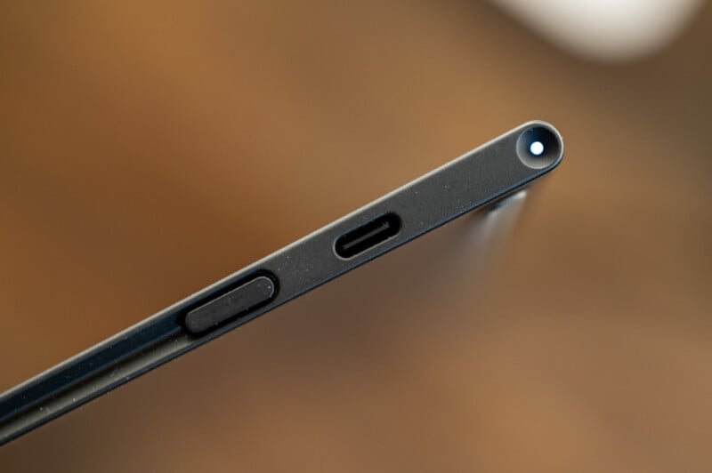 A close-up image of the edge of a black electronic device, possibly a tablet or thin laptop. The edge features a USB-C port, a small button, and another small circular port. The background is blurred, highlighting the sharp focus on the device's edge.