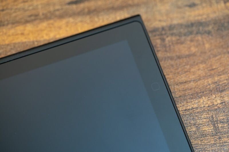 Close-up of the corner of an electronic device, possibly a tablet or phone, showing part of its screen and a thin black frame. The background surface is wooden with a slightly rough texture and visible grain patterns.