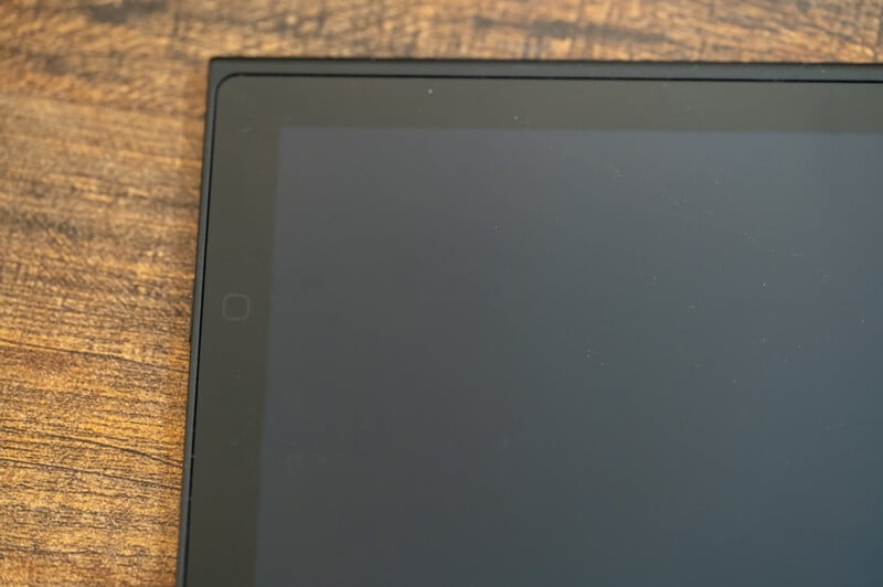 Close-up of a black tablet with the screen turned off, positioned on a wooden surface. Only part of the screen and the surrounding bezel are visible in the frame.
