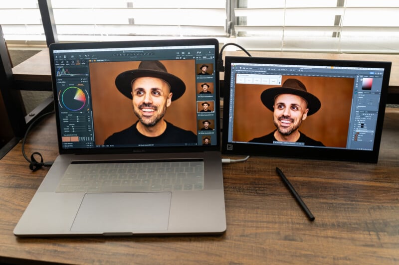 A laptop and an external monitor display an image editing application with the same photo of a smiling person wearing a hat. The workspace is on a wooden desk with a stylus pen resting nearby. The blinds in the background are partially open, letting in some light.