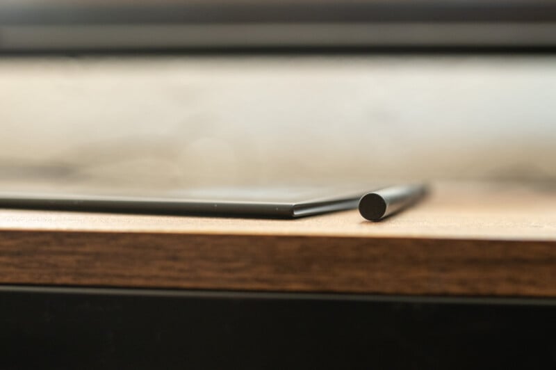 A close-up side view of a modern tablet laid flat on a wooden surface, with its black stylus pen placed next to it horizontally. The background is blurred, highlighting the tablet and pen's sleek design.