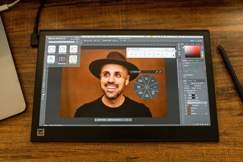 A tablet screen displays a photo editing software interface with an image of a smiling person wearing a brown hat on the screen. The tablet is placed on a wooden desk with a stylus and part of a laptop visible nearby.