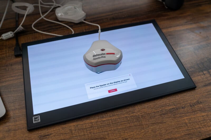 A Spyder Pro color calibration tool is placed on a flat monitor screen lying on a wooden desk. The monitor displays a prompt instructing the user to place the Spyder on the indicated area. Cables and a wall charger are visible in the background.