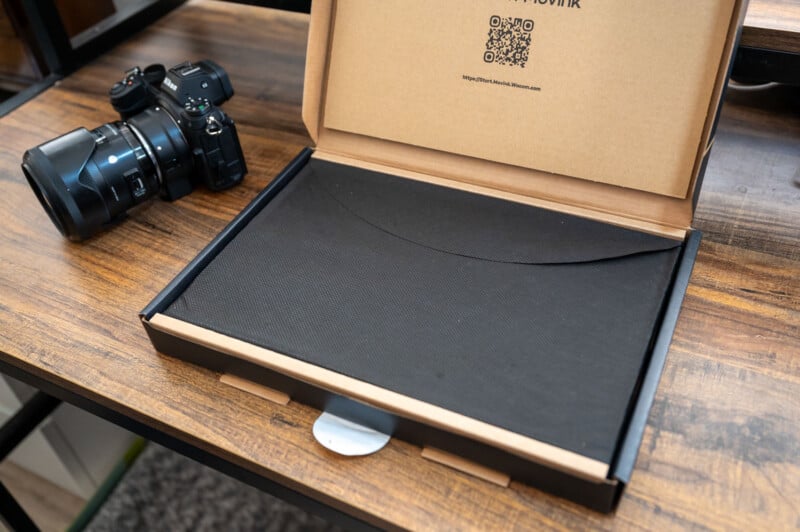 An open cardboard box with a black envelope inside rests on a wooden table. Next to the box is a digital camera with a lens cap attached and an additional lens lying beside it. The background includes parts of a table and a shelf.