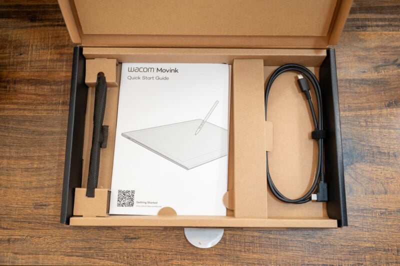 A Wacom Movink package contains a pen tablet, quick start guide, black pen with a textured grip, and a USB cable. The items are neatly arranged in a cardboard box with a wood-patterned background. A QR code for getting started is visible on the guide.