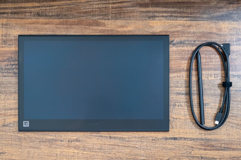 A Wacom drawing tablet lies flat on a wooden surface. Next to it is a black HDMI cable, neatly coiled and secured with a Velcro strap. The tablet's screen is off, displaying a black surface with a small logo in the corner.