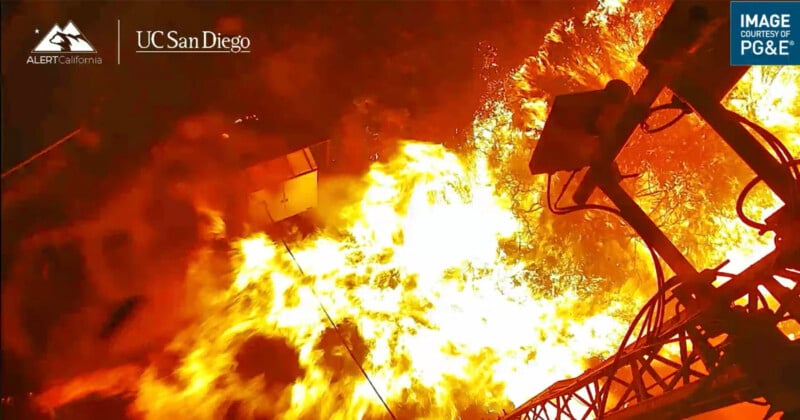 A wildfire rages with intense yellow flames engulfing the scene, partially surrounding an outdoor setup with technological equipment. The fire's bright light contrasts against the dark background. Logos for ALERTCalifornia, UC San Diego, and PG&E are visible.