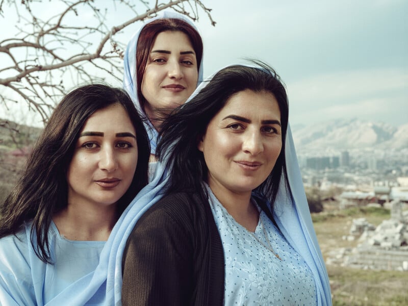 Three women are standing outdoors against a scenic backdrop of hills and a cityscape. They are smiling and wearing light blue tops and shawls. A leafless tree branch is visible in the top left corner of the image. The sky is partly cloudy.