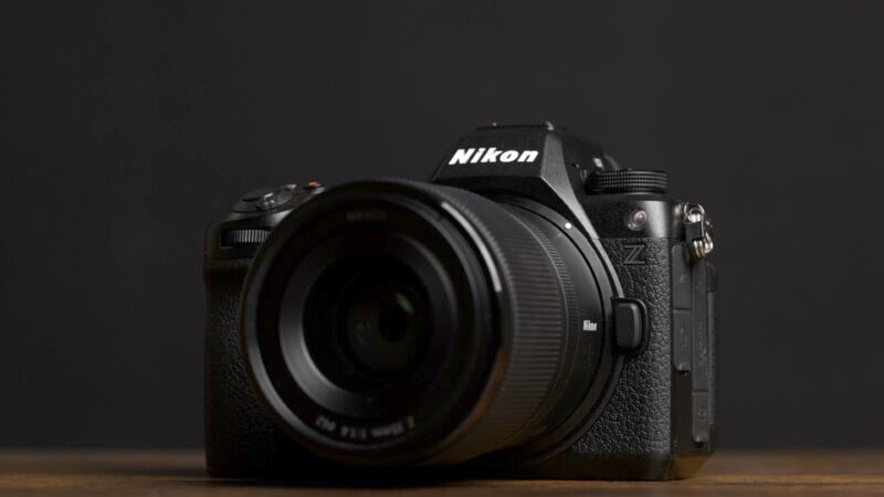 A Nikon Z series camera with a lens attached, placed on a wooden surface against a black background. The Nikon logo is visible on the camera body.