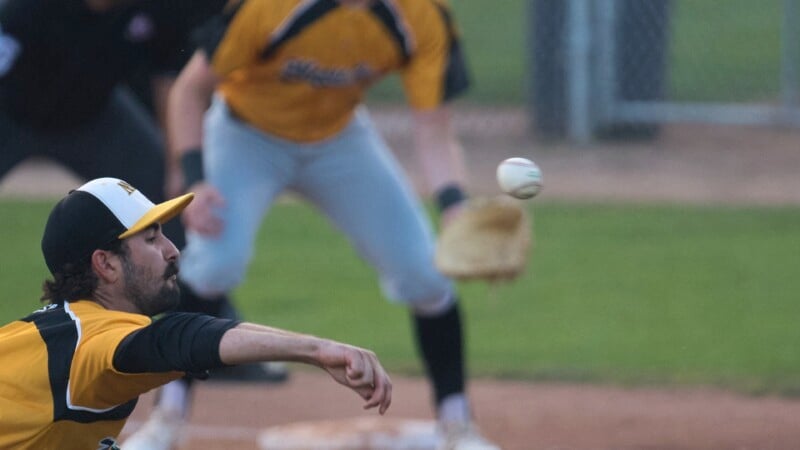 A baseball pitcher in a yellow and black uniform is in mid-throw while a player in the background, also wearing yellow and black, stands ready with a glove. The baseball is visible in the air. The field and a fence are in the background.