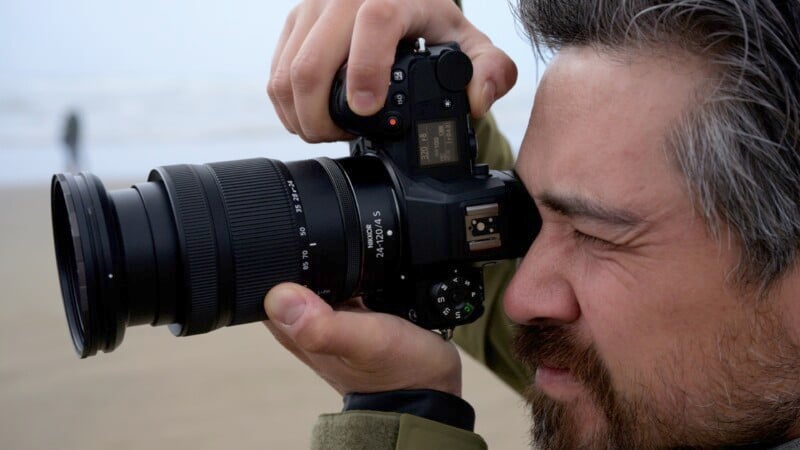 A close-up of a person with grey-streaked hair taking a photo using a DSLR camera with a large lens, held up to their eye. The person appears focused, with a blurry beach and another person in the distant background.