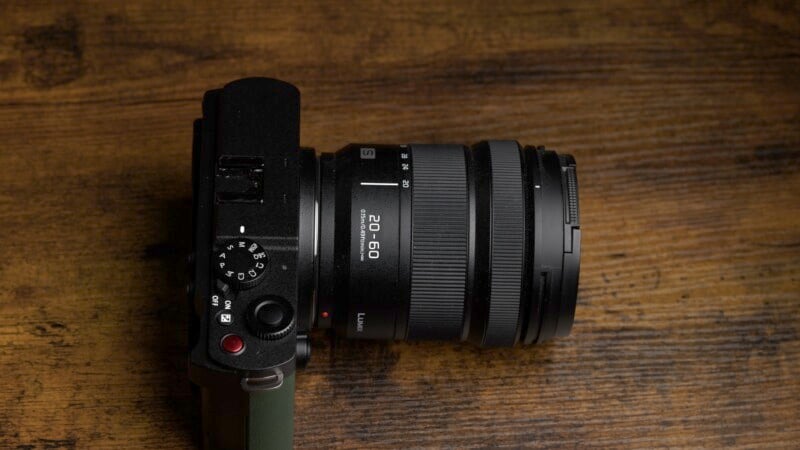 A black digital camera with a lens labeled "20-60" placed on a brown wooden surface. The camera has various buttons and dials, including a red button and a green strap on the left side. The image is taken from an overhead angle.