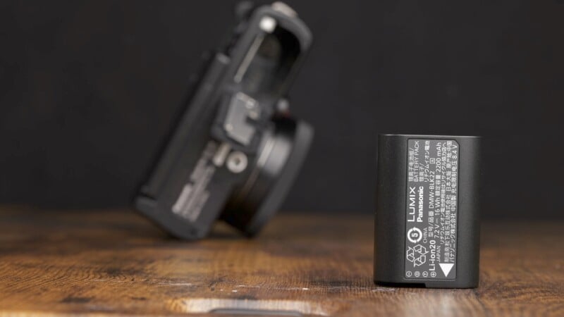 A close-up of a Lumix rechargeable lithium-ion battery resting on a wooden surface, with a blurred view of a Lumix camera in the background. The battery has labels and specifications printed on it, and the image is taken in a dimly lit setting.