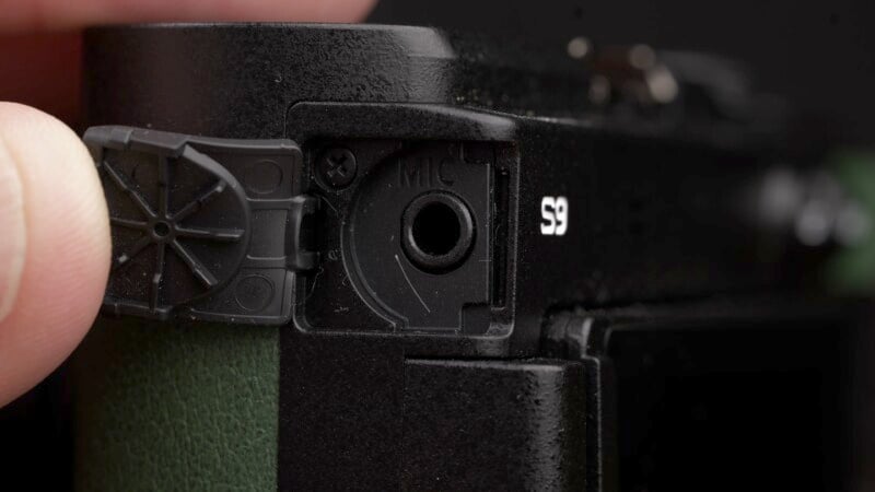 Close-up of a hand opening the microphone jack cover on a black camera. The cover is partially opened, revealing the microphone input labeled "MIC." The camera body features a text marking "S9" near the top-right corner. The background is out of focus.