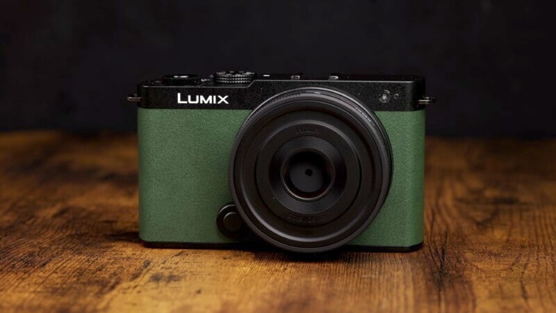 A green Lumix camera with a black lens sits on a wooden surface against a dark background. The camera has a textured finish and a minimalist design, featuring simple control buttons and a compact build.