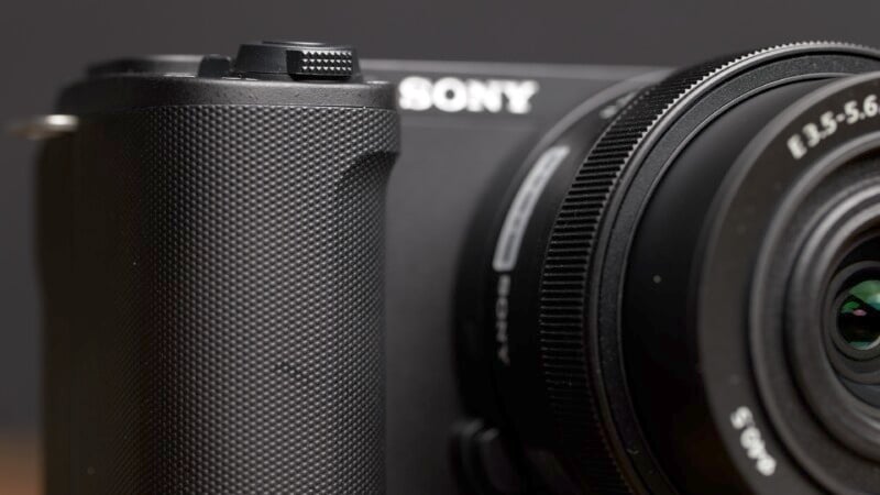 Close-up view of a black Sony camera, focusing on the textured grip, with part of the lens visible on the right. The Sony logo is partially visible at the top. The background is blurred.