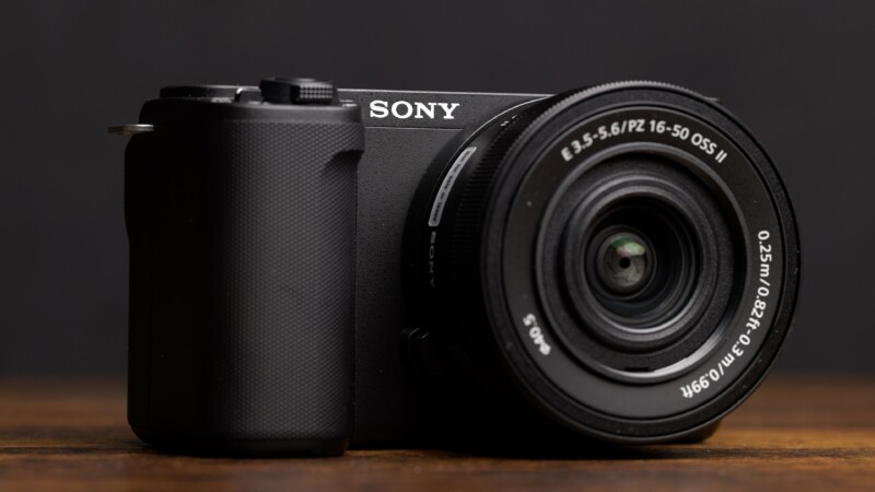 Close-up image of a Sony mirrorless camera with an attached zoom lens resting on a wooden surface. The camera body is black with a textured grip on the left side. The lens has detailed markings and text visible on its circumference, focusing on the 16-50mm range.