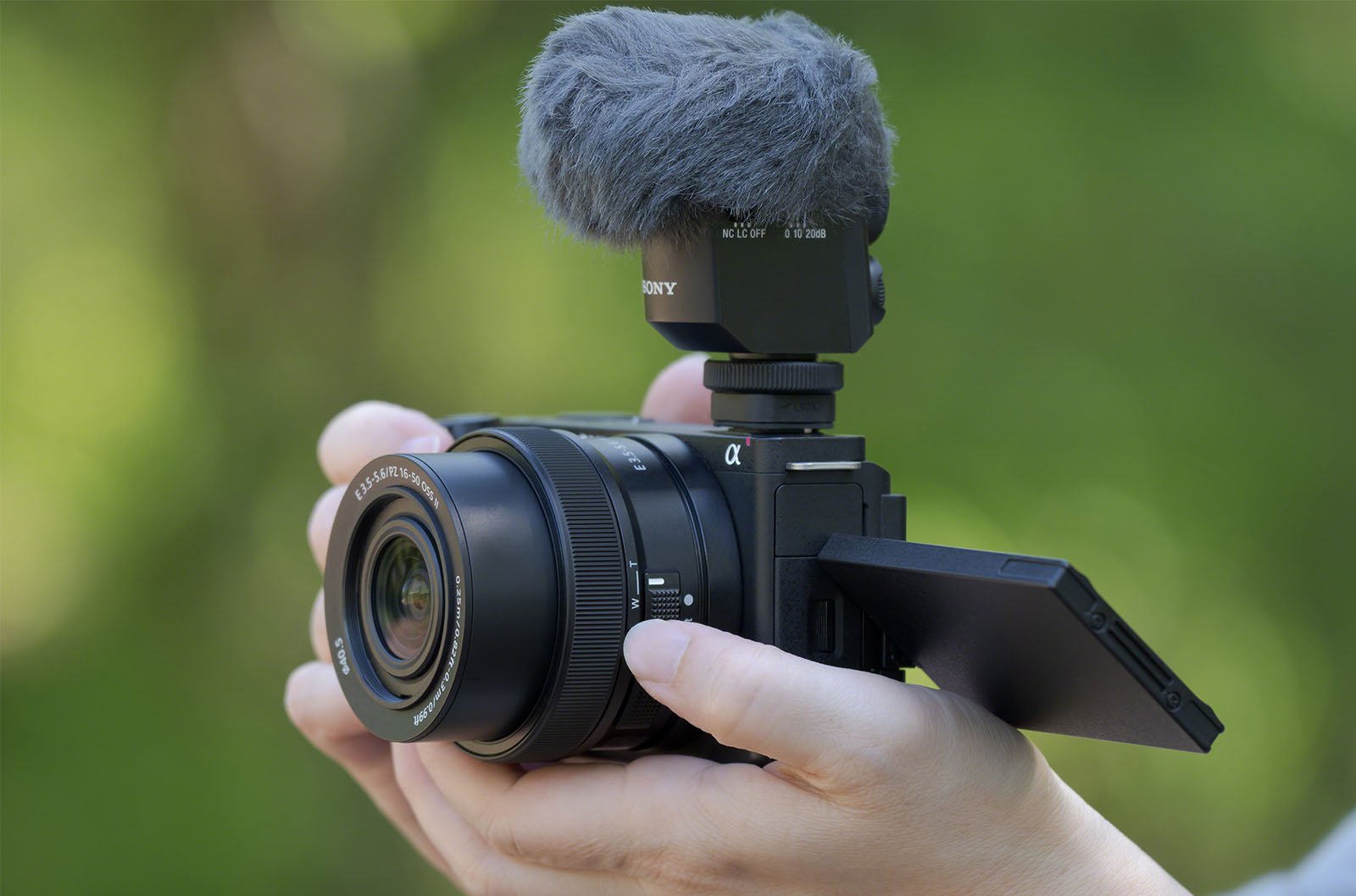 A person is holding a digital camera with a large lens and a flip-out screen. The camera is equipped with an external microphone that has a fluffy windscreen. The background is blurred, highlighting green foliage.