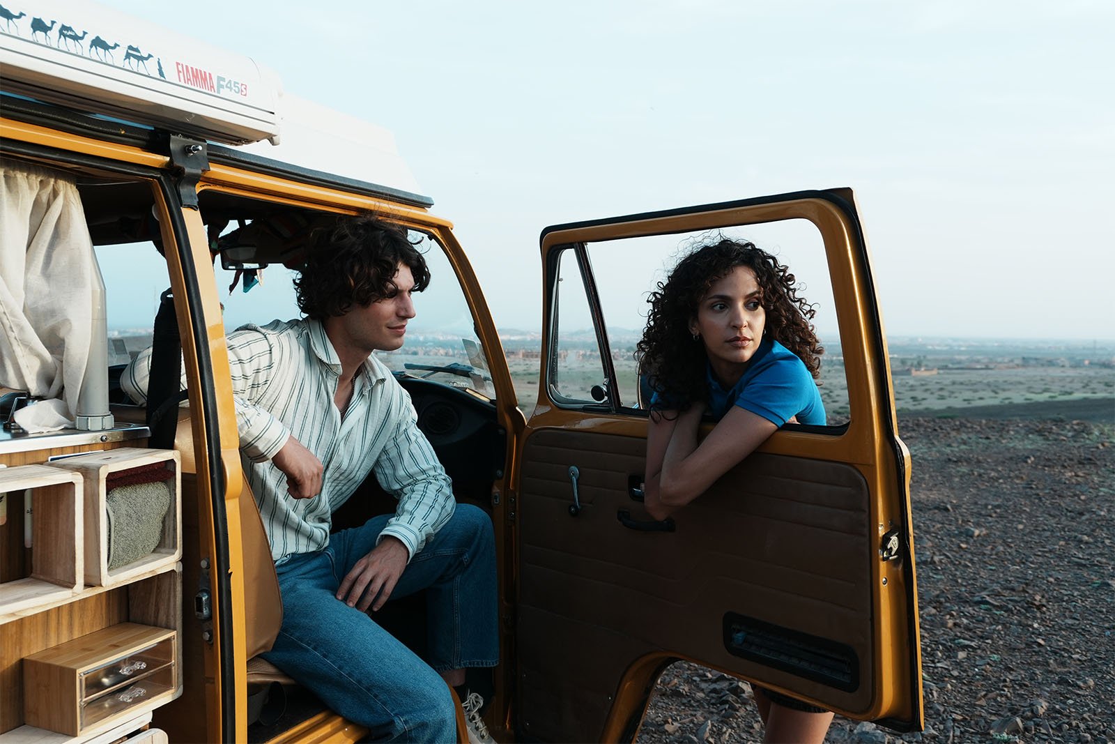 A young man and woman with curly hair lean against a yellow van in a desert landscape. The man sits inside the van, while the woman leans on the open door, gazing into the distance. The interior of the van shows shelves with belongings.