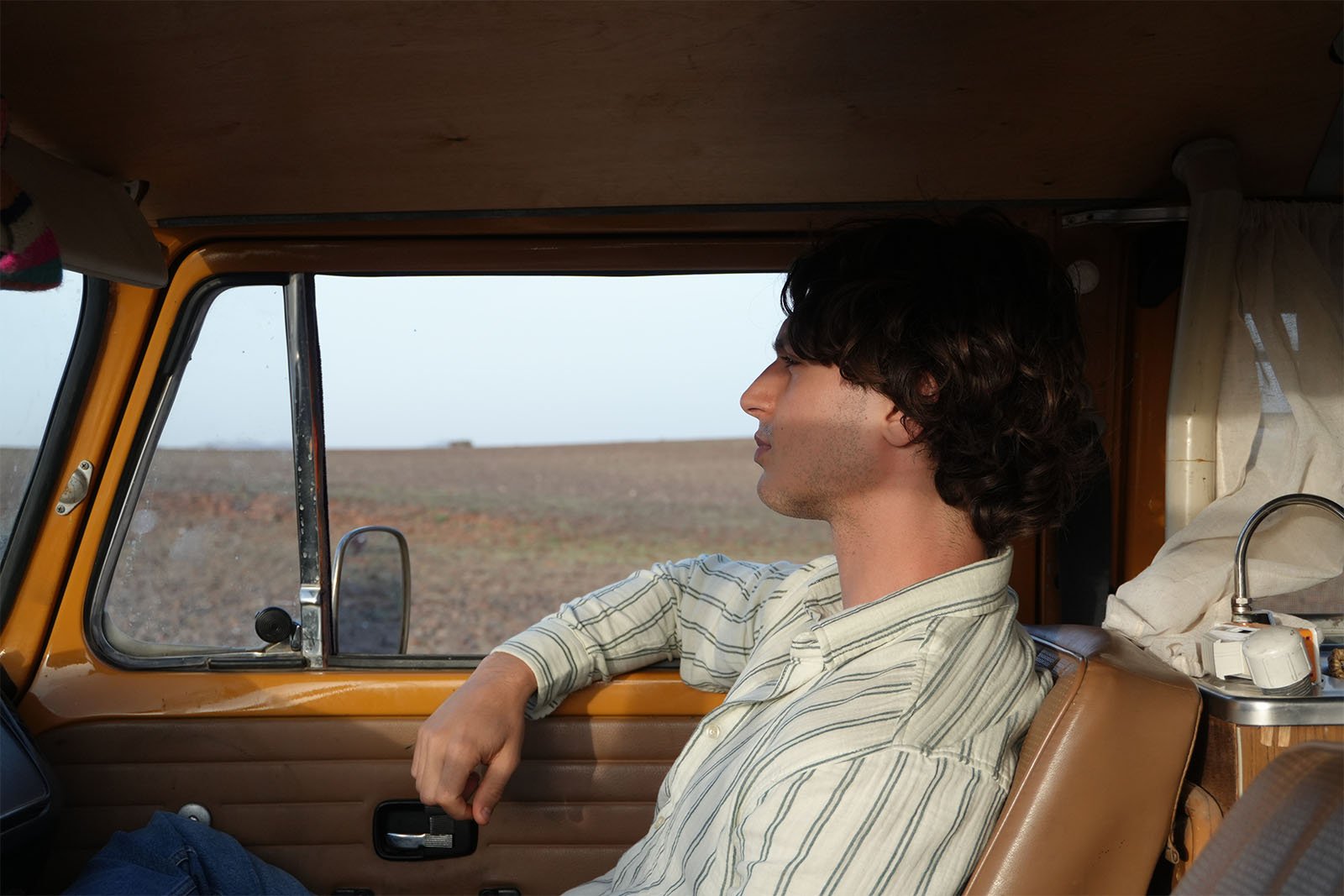 A person with shoulder-length brown hair, wearing a striped shirt, sits in the driver's seat of a vintage car gazing out the window. The background shows a vast, barren landscape with clear skies. The interior of the car has a rustic, wooden vibe.