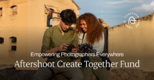 A man and a woman smile and look at the screens of the cameras they are holding. They are outdoors with a building in the background. Text reads, "Empowering Photographers Everywhere. Aftershoot Create Together Fund." The Aftershoot logo is in the top right corner.
