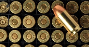 Close-up of a tray filled with .45 Auto pistol cartridges, showing the brass casing with "45 AUTO" markings. One cartridge is positioned horizontally above the others, displaying its full length and copper jacketed bullet. The background is black.