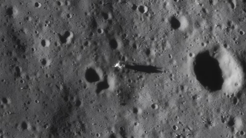 A high-resolution image of the Moon's surface showing a large number of craters. One prominent crater on the right side of the image casts a dark shadow. In the center, a bright object with a long shadow stretches across the lunar terrain.