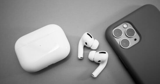 A pair of AirPods lie next to their white charging case on a gray surface. Beside them is a smartphone with a black case, featuring multiple camera lenses on the back.
