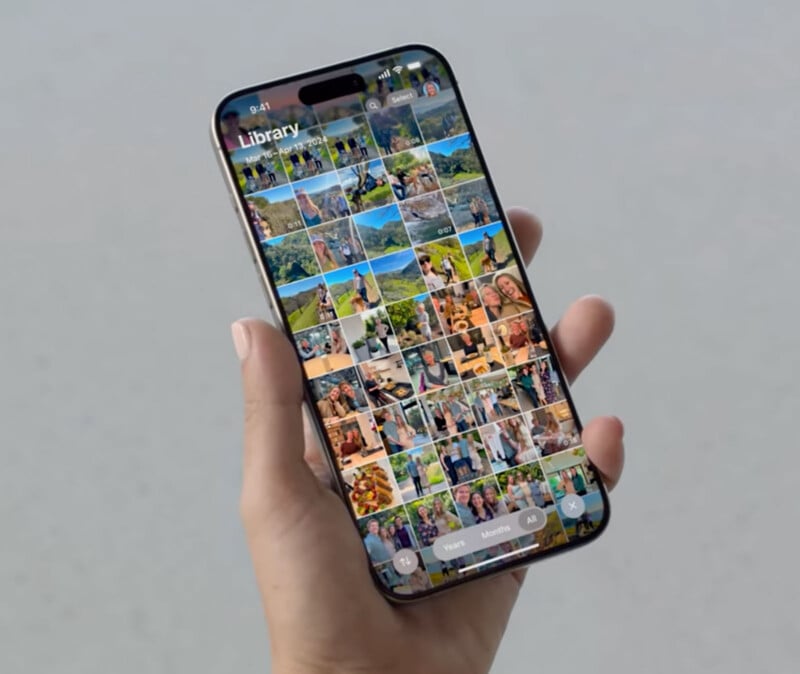 A person's hand holds a smartphone displaying a photo library screen filled with various thumbnail images. The phone's screen shows multiple rows of colorful photos, suggesting various moments and events. The background is a plain, light surface.