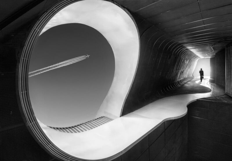 A black-and-white image of a person walking through a futuristic, wavy architectural tunnel. The tunnel curves organically, opening to the sky on the left side where a contrail from a passing airplane is visible. The person is silhouetted against the bright light.