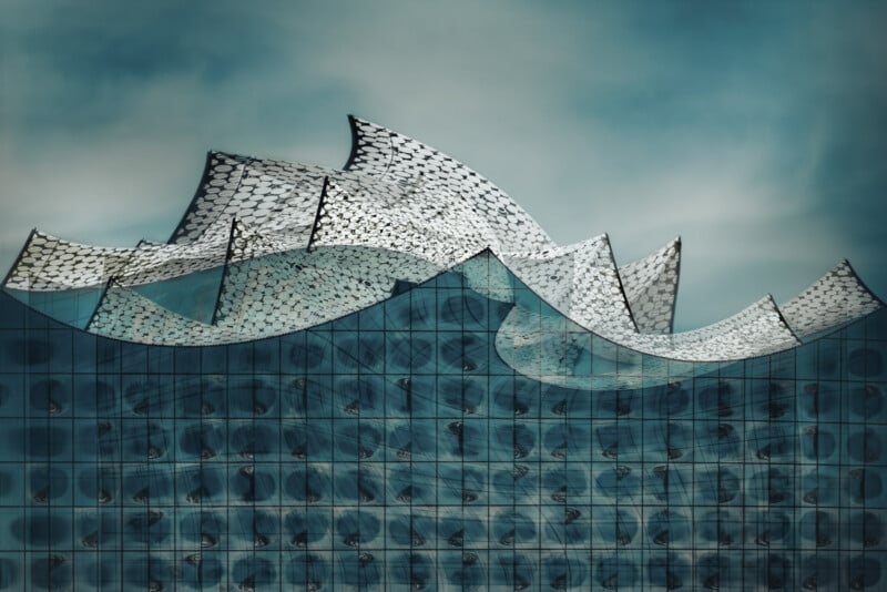 A modern building with a glass facade features a wavy, sail-like roof structure. The intricate roof pattern and the reflective glass create an artistic, dynamic appearance against a blue sky backdrop.