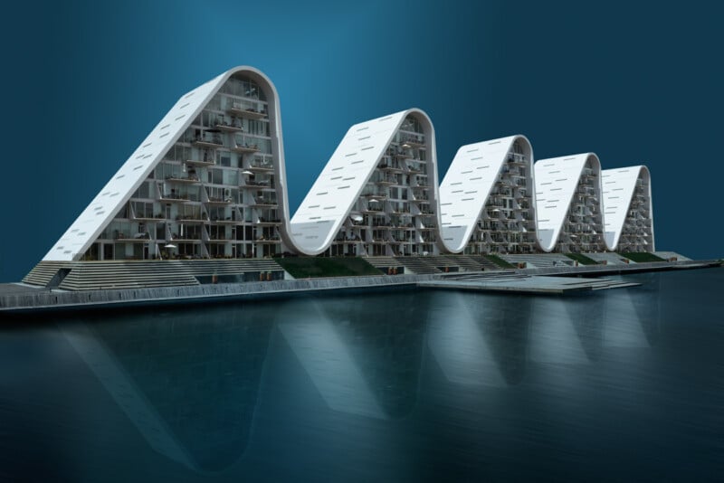 A row of modern, wave-shaped apartment buildings with white facades stand along a calm waterfront. The unique architecture features curved roofs and large windows. The structures are mirrored in the still water below, creating a symmetrical reflection.