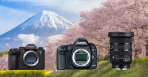 Three camera items are arranged in front of a background featuring cherry blossoms and a snow-capped mountain. From left to right: Sony Alpha camera body, Canon EOS 5D Mark III camera body, and a Sigma zoom lens. The scene portrays a serene, scenic landscape.
