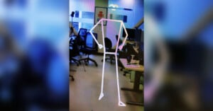 A skeletal representation of a humanoid figure is shown superimposed on a blurred background of an office or lab environment. The background includes office chairs, desks, and computer screens. The skeletal figure is made up of white lines depicting basic body structure.