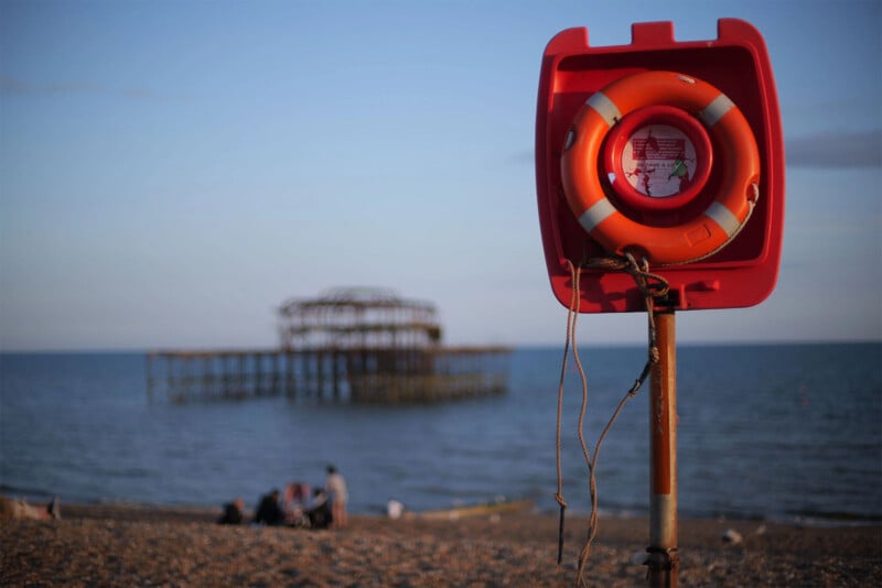 A bright orange life preserver is mounted on a red post on the shore, with an old, dilapidated pier structure in the background extending into the blue sea. The sky is clear, and a few people are visible sitting on the beach near the water's edge.